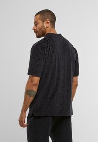 Short-sleeve black shirt with a textured geometric pattern, featuring a collar and relaxed fit. Material appears soft and slightly plush.