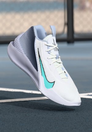 Chaussure de sport Nike blanche et grise avec un logo bleu sarcelle, lacets noués, flottant au-dessus d'une surface de court de tennis bleu près d'une ligne blanche.