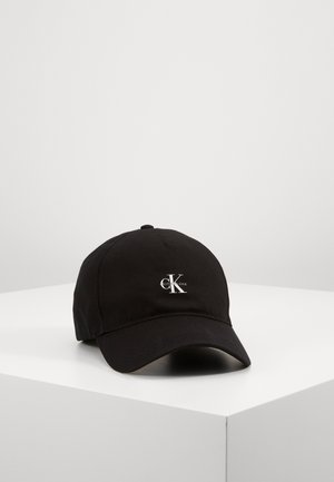 Czarna czapka z daszkiem w stylu baseballowym z zakrzywionym daszkiem i małym białym logo "CK Calvin Klein Jeans" z przodu, położona na białej powierzchni.