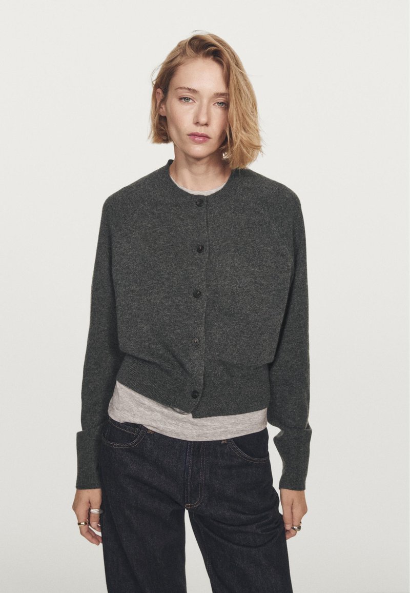 Koolgrijze, cropped cardigan gemaakt van zacht materiaal, met een ronde halslijn, knopenfront en lange mouwen over een grijze T-shirt.