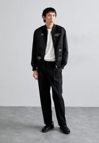 Giacca bomber nera con pattern in rilievo, maglietta bianca a girocollo, pantaloni neri e sneakers nere. Design semplice, senza hardware visibile.