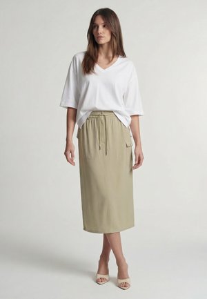 Frau steht und trägt ein lockeres weißes V-Ausschnitt-T-Shirt, einen beigen Midirock mit Kordelzug und Tasche sowie beige offene Sandalen mit Absatz.