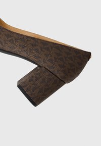 Smeđe cipele s potpeticom s teksturiranim uzorkom, kvadratnom blok-petom i glatkim unutrašnjim dijelom. Imaju nisku kroja s šiljastim prstima.