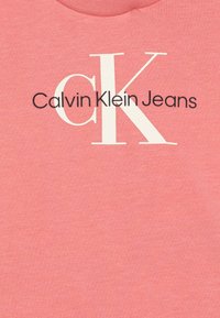 Růžové bavlněné tričko s bílým logem "CK" a textem "Calvin Klein Jeans" v černé barvě, s pruhovaným výstřihem a hladkou texturou.