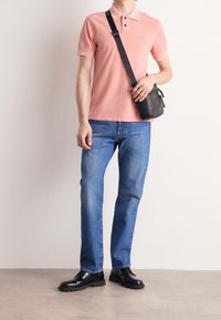 Polo shirt rose clair avec un col et deux boutons, associé à un jean bleu et des chaussures noires. Un petit sac à bandoulière noir est porté sur l'épaule.