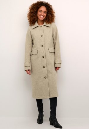Beige, strukturierter Mantel mit Kragen, langen Ärmeln, Frontknöpfen und zwei seitlichen Taschen, getragen über einer dunklen Jeans und Stiefeletten.