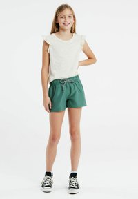 Chemise beige clair à manches volantées, associée à un short vert à cordon de serrage et des baskets montantes noires. Fond blanc.