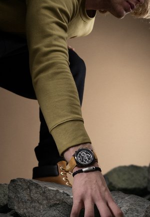 Montre chronographe noire avec un grand cadran rond, un bracelet en cuir et des accents argentés. Sweat à capuche vert olive ; bottes robustes ; surface rocheuse.