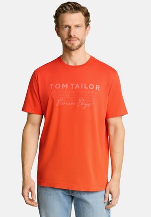 Mann mit hellbraunen Haaren und Bart, trägt ein oranges "Tom Tailor" T-Shirt und hellblaue Jeans, steht vor weißem Hintergrund.