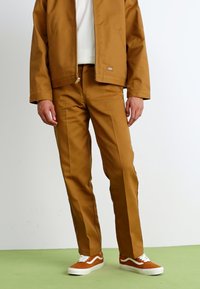 Brown waterproof jacket and matching pants, made of smooth fabric, with a relaxed fit. White sweatshirt underneath and orange sneakers.