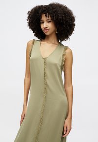 Oliwkowa sukienka bez rękawów z dekoltem w kształcie litery V, z frędzlami wzdłuż krawędzi i gładkiego, lekkiego materiału.