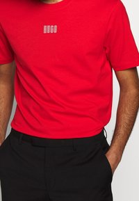 Mand iført en lys rød T-shirt med et lille hvidt "HUGO" logo på brystet, kombineret med sorte bukser, hænderne i lommerne.