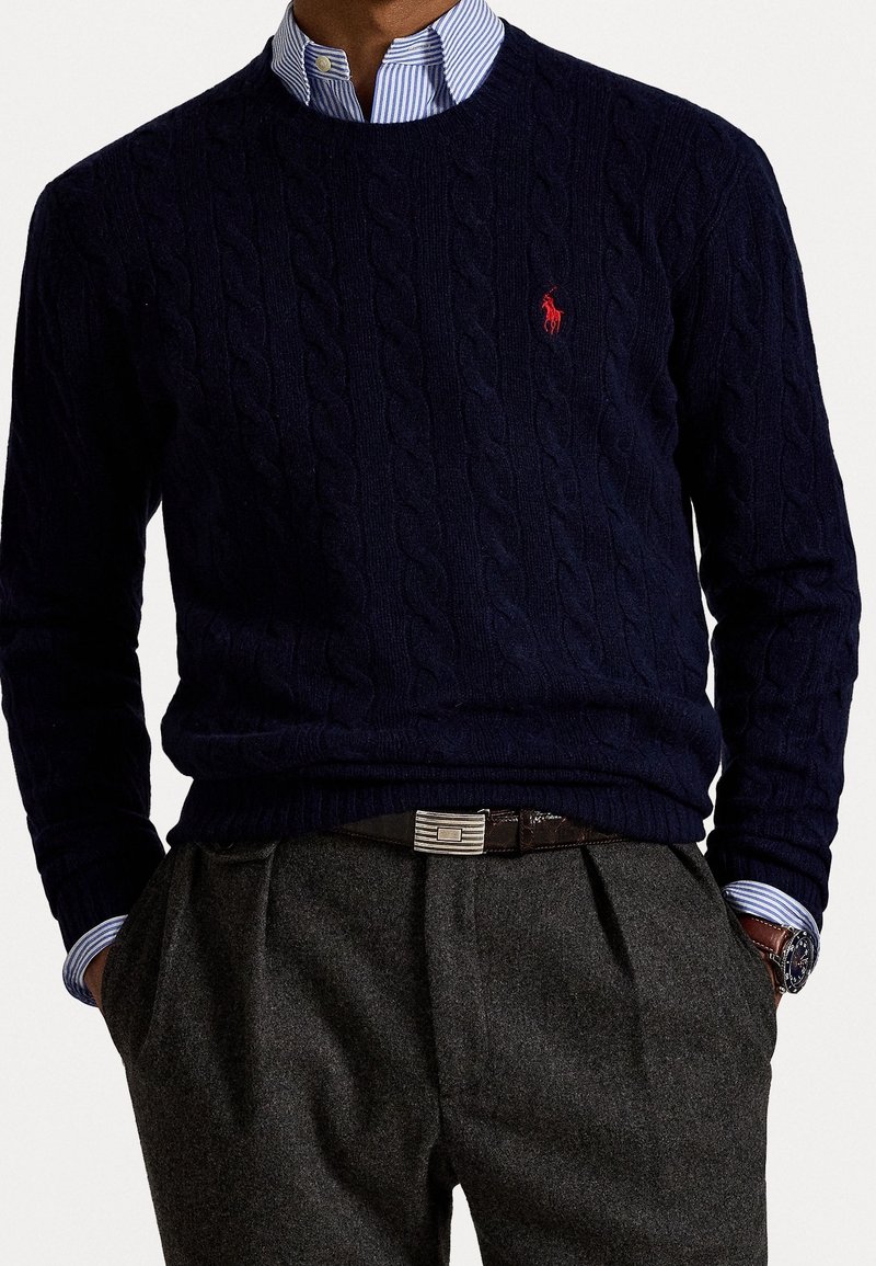 Homme portant un pull en maille torsadée bleu marine avec un logo rouge, une chemise rayée bleue et blanche, un pantalon gris, une ceinture noire et une montre-bracelet.