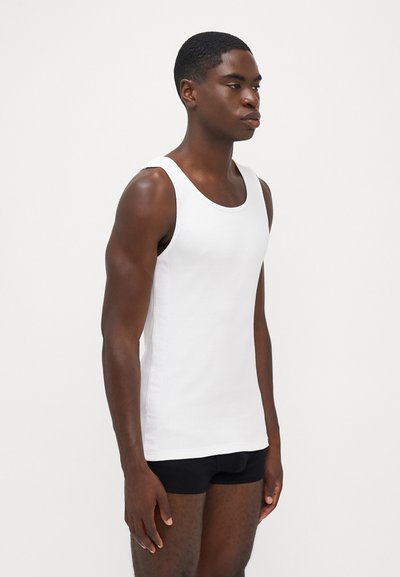 Tricou alb cu dungi, cu bretele late, formă ajustată și textură moale. Purtați deasupra unui şort sport negru, purtat de un model masculin.