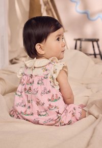 Salopetă roz din bumbac cu modele de iepuraș și flori, având un decolteu cu volane și detaliu cu nasturi în spate, purtată de un copil mic.