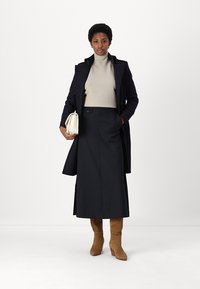 Tengerész stílusú trench coat, bézs garbó, fekete szoknya, világosbarna térd fölé érő csizma és egy fehér kézitáska semleges háttér előtt.