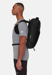 Mochila negra con forma redondeada, tela texturizada, correas ajustables y detalles en naranja. La lleva puesta una persona con una camiseta gris y pantalones oscuros.