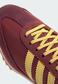Maron sportssko med gule snørebånd, med en tekstureret stof- og ruskindsoverdel, zigzag-stribede detaljer og en gummisål.