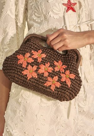 Mână ținând o geantă maro țesută cu aplicații de flori portocalii, purtând o rochie albă brodată cu flori și o broșă în formă de stea de mare.