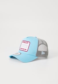 Gorra trucker de malla azul con panel frontal de tela, parche de logo en blanco y rojo, visera plana y parte trasera ajustable para personalización del tamaño.