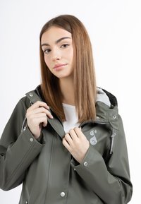 Jachetă verde măslinie rezistentă la apă cu glugă, accesorii argintii, manșete elastice, închidere cu șnur și un logo pe față.