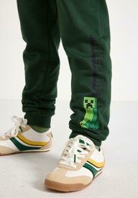 Pantaloni della tuta verdi con logo 'Minecraft' nero e grafica del Creeper. Scarpe beige e bianche con strisce verdi e gialle. Texture liscia, design casual.