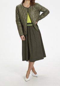 Chaqueta acolchada de color verde oliva con cuello redondo y botones a presión, combinada con una blusa de color verde brillante y una falda larga oscura. Tacones blancos.