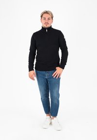 Maglione nero lavorato a maglia con zip e colletto alto, abbinato a jeans azzurri aderenti e sneakers bianche, in piedi davanti a uno sfondo bianco.