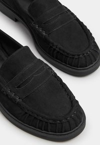Mocassini in suede nera con dettagli cuciti e punta tonda. Presentano una cucitura rialzata lungo la parte superiore e una suola flessibile e texture.