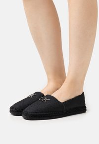 Sorte slip-on sko med tekstureret stof, der har et logo-akcent og en flettet jutesål for ekstra detalje. Designet fremhæver komfort og stil.