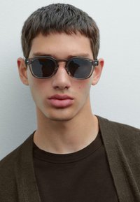 Round sunglasses with dark lenses, transparent gray frames, highlighting a minimalist design. The wearer has short hair and neutral-toned clothing.