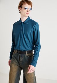 Teal ribbed long-sleeve shirt with a collar and button-down front, paired with high-waisted gradient denim jeans and a black belt.
