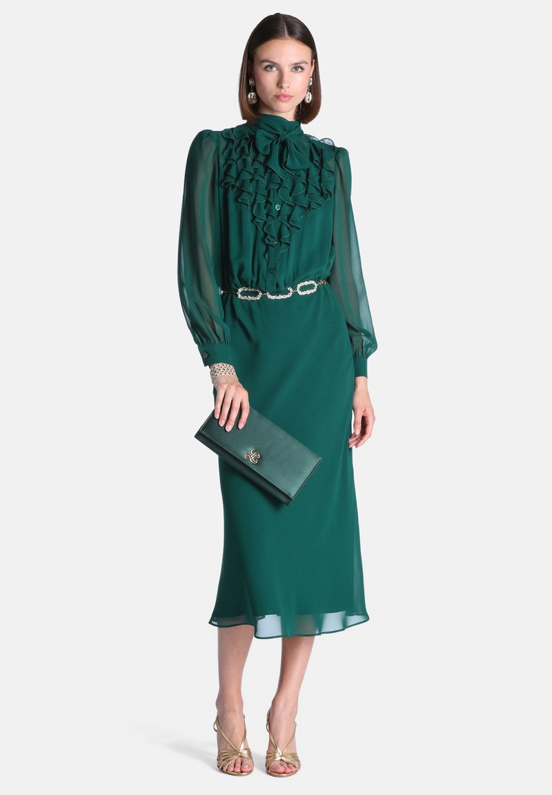 Femme portant un chemisier à manches longues vert foncé à volants et une jupe midi assortie, tenant une pochette verte et portant des sandales et des bijoux en or.