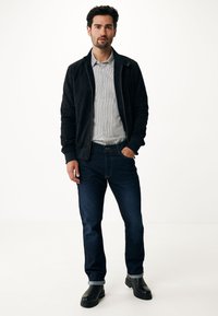 Chaqueta bomber negra sobre una camisa de rayas clara, combinada con jeans azul oscuro y botas negras. Tejido suave, diseño ajustado, puños enrollados.