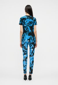 Γυναίκα με μακριά σκουρόχρωμα μαλλιά που στέκεται, φοράει μαύρη και μπλε κοντομάνικη κορυφή και παντελόνι με στροβιλιστό μοτίβο και μαύρα ψηλοτάκουνα παπούτσια.