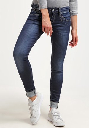 Person, die eng anliegende dunkelblaue hochgekrempelte Jeans und graue Sneakers mit Klettverschluss trägt, steht mit übereinandergeschlagenen Beinen.