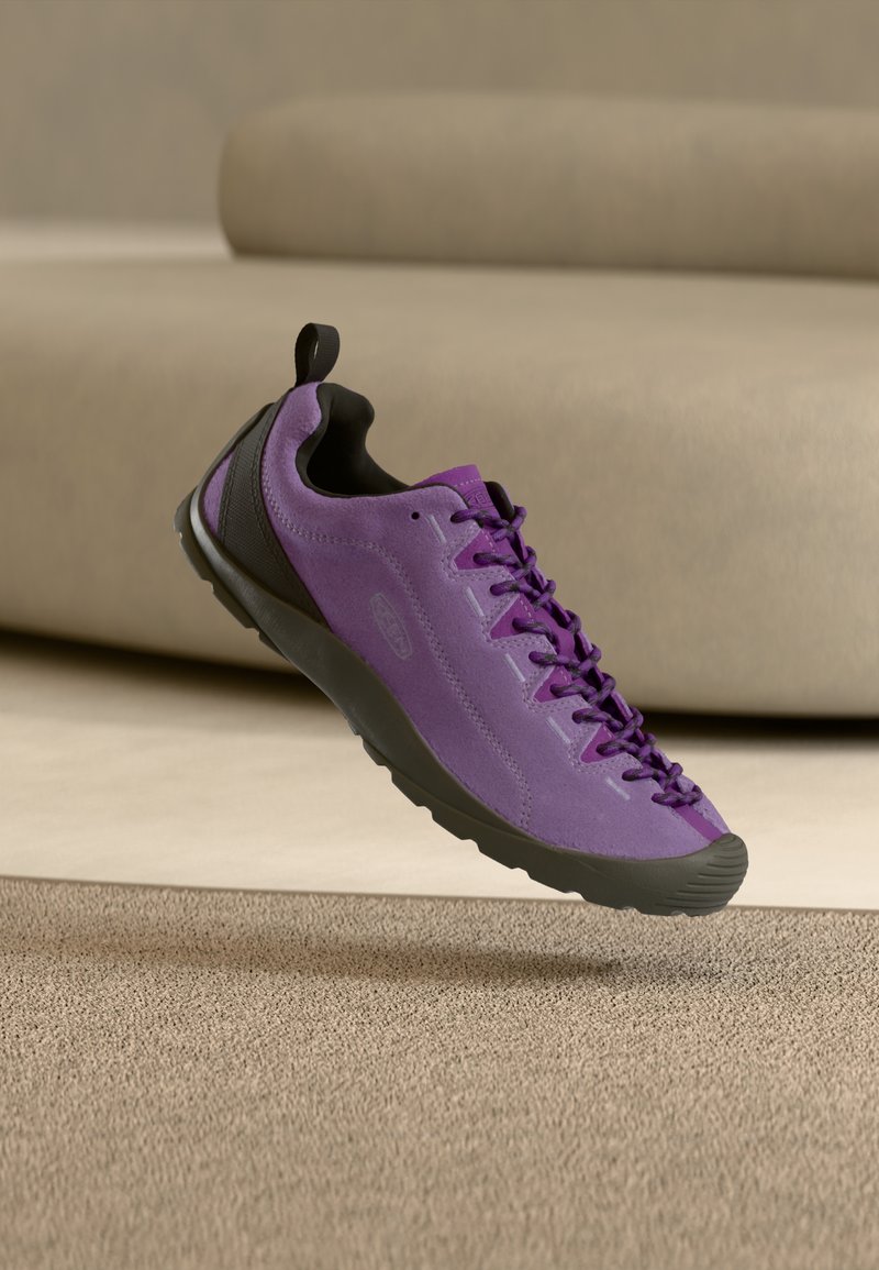 Μωβ πάνινο παπούτσι από καστόρι με μαύρη σόλα και κορδόνια, αιωρούμενο πάνω από μπεζ χαλί με ένα μπεζ καναπέ στο παρασκήνιο.