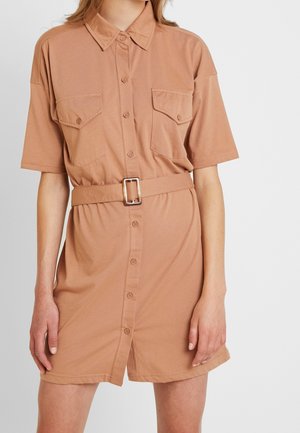 Femme portant une robe-chemise à manches courtes beige avec des poches poitrine et une ceinture assortie avec une boucle rectangulaire.