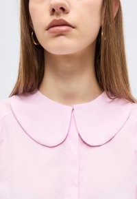 Jeune femme aux cheveux lisses châtain clair portant des boucles d'oreilles créoles en or et un chemisier rose clair avec un col arrondi, sur un fond uni.