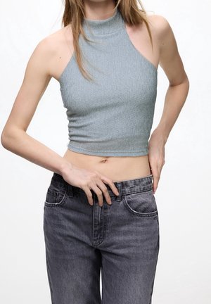 Femme portant un crop top côtelé bleu clair à col halter et un jean noir délavé taille haute, ajustant la taille avec ses mains.