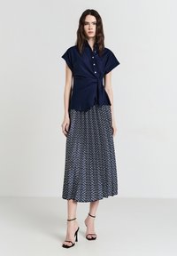 Donna che indossa una blusa blu navy con maniche corte e bottoni, abbinata a una gonna midi a pattern blu e bianco, con sandali neri con tacco alto.