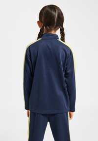 Marinblå sweatshirt med dragkedja i halsen och långa ärmar, med ljusgula sidoränder. Mjukt, texturerat tyg; avslappnad passform.