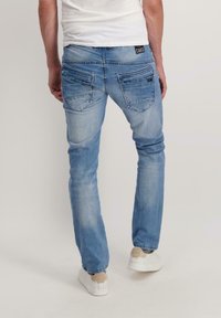 Persoon in lichtblauwe denim jeans met flapzakken aan de achterkant en witte sneakers, staand tegen een effen achtergrond.