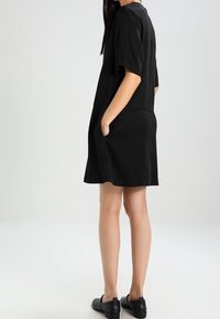 Robe noire à manches courtes avec une coupe ample, poches des deux côtés et une texture lisse. Portée avec des chaussures plates noires.