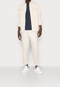 Camisa de botones de color beige claro usada sobre una camiseta azul marino con estampado, combinada con pantalones beige claro y zapatillas blancas. Textura de tela suave.