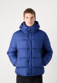 Mėlynas pūkinis švarkas su aukštu apykaklę, horizontaliomis siūlėmis, priekiniais mygtukais ir reguliuojama sutraukimo kapišonu. Lengvas, elegantiškas dizainas.