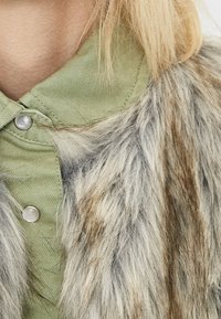 Chemise verte à col avec des boutons-pression argentés, superposée avec de la fourrure synthétique présentant des tonalités grises et brunes, mettant en avant des textures variées.