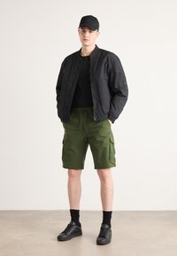 Svart bomberjakke over en svart T-skjorte, sammen med grønne cargoshorts. Sorte sneakers og caps fullføre antrekket. Minimalistisk design.