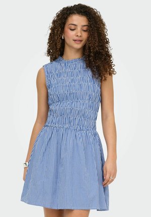 Jeune femme aux cheveux bouclés portant une robe sans manches à carreaux bleu et blanc, debout les yeux fermés devant un fond uni.