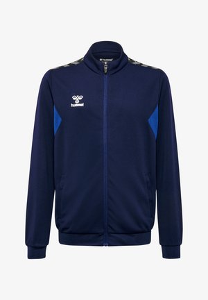 Mørkeblå lynlåstræningsjakke i tekstureret stof, med blå sideaccenter og et mønster på skulderen. Hummel-logo foran.